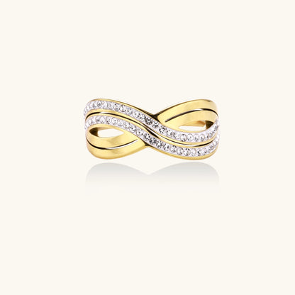 Gold-plated stainless steel ring with crossed infinity bands adorned with sparkling rhinestones against cream background.